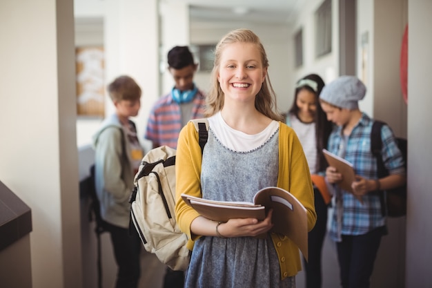 Porträt des lächelnden Schulmädchens, das mit Notizbuch im Korridor steht