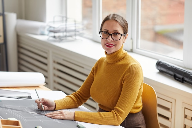 Porträt des lächelnden weiblichen Architekten, der Blaupausen und Pläne zeichnet, während er am Schreibtisch im Büro arbeitet, und