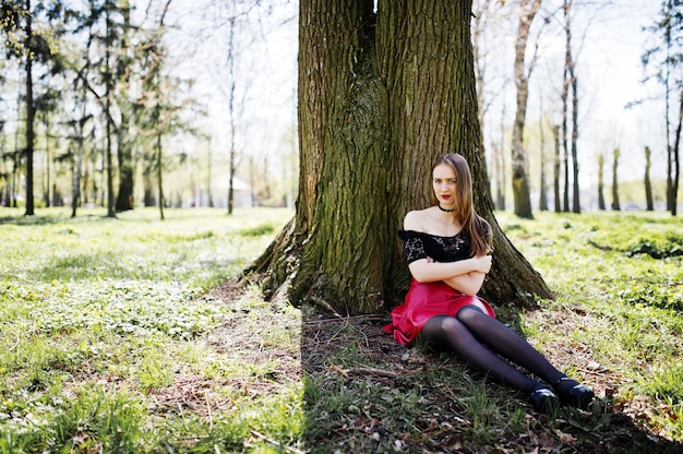 Porträt des Mädchens mit hellem bilden mit den roten Lippen, schwarzer Chokerhalskette auf ihrem Hals und rotem Lederrock, die nahe Baum am Park sitzen.