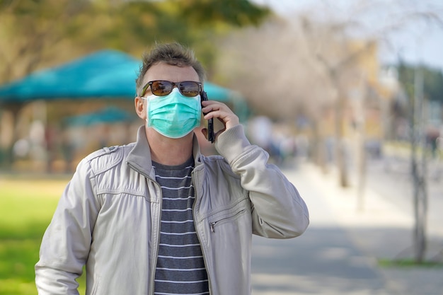 Porträt des Mannes auf der Straße in einer medizinischen Maske und spricht am Telefon. Attraktives unglückliches Modell mit Grippe im Freien. Unglücklicher Mann mit Grippe im Freien.