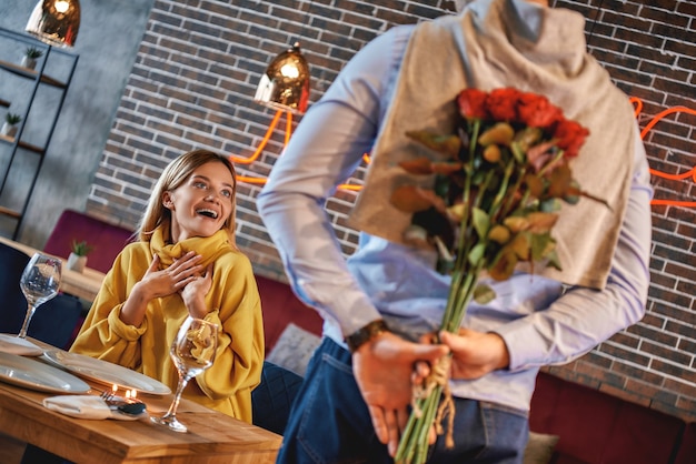 Porträt des Mannes, der mit einem Strauß Rosen im Restaurant steht. Frau freut sich, ihr Date zu sehen. Romantisches Dinner-Konzept. Horizontale Aufnahme