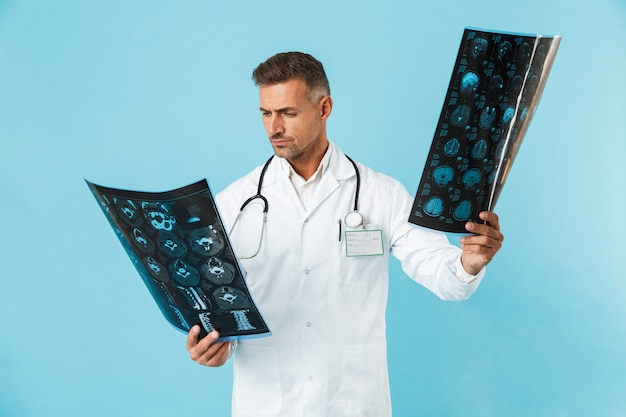 Porträt des medizinischen Arbeiters 30s mit Stethoskop, das Röntgenbilder hält, lokalisiert über blauer Wand