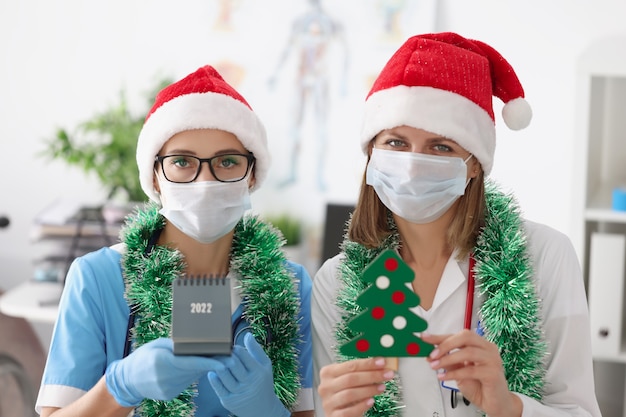 Porträt des medizinischen Personals trifft neues Jahr in Gesichtsmasken, festlichen Hüten und Dekor. Arzt und Krankenschwester an der Rezeption. Medizin, Gesundheitswesen, Pandemie, Quarantäne, Konzept zur Verhinderung der Virusverbreitung