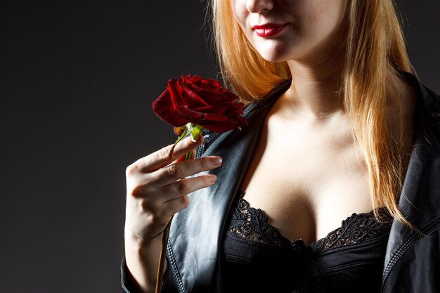 Porträt des Modells der jungen Frau im roten Kleid, das rote Rosenblume in der Hand über dunklem Hintergrund im Fotostudio hält Schönheits- und Modekonzept