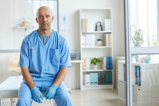 Porträt des modernen Mannes, der blaue Uniform trägt, die im Krankenhaus arbeitet, das in seinem Büro sitzt