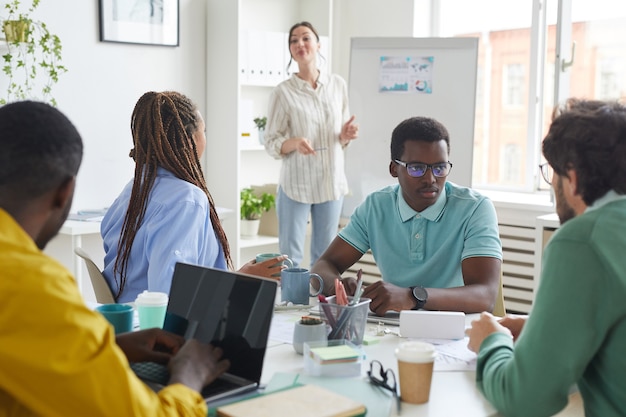 Porträt des multiethnischen Geschäftsteams, das Projekt während des Sitzens am Tisch im Konferenzraum mit dem Manager diskutiert, der durch Whiteboard steht