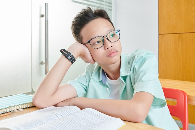 Porträt des nachdenklich gelangweilten vietnamesischen Schuljungen in den Gläsern, die auf Hand lehnen und durch Fenster schauen