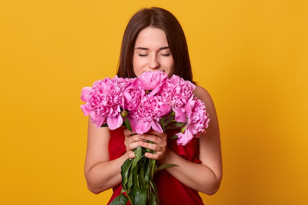 Porträt des netten jungen brünetten Mädchens, das Strauß der rosa Pfingstrosen gegen gelbe Studiowand hält, Kleid trägt, Blumen riecht