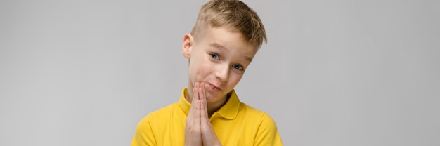 Porträt des netten kleinen blonden kaukasischen Jungen im gelben T-Shirt, das um Vergebung auf grauem Hintergrund bittend hofft