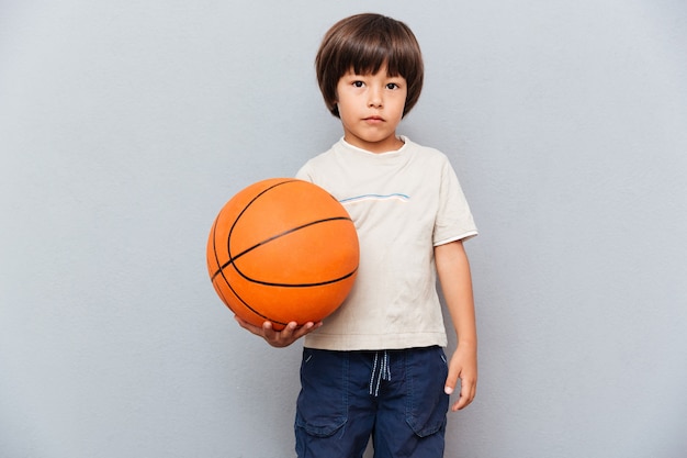 Porträt des netten kleinen Jungen, der Basketballball steht und hält