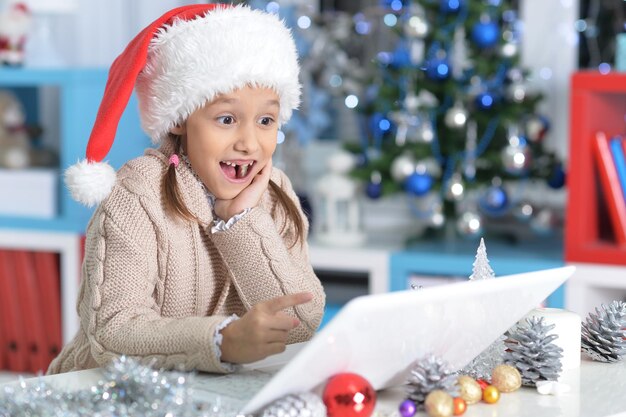 Porträt des netten kleinen Mädchens in der Weihnachtsmütze mit Laptop