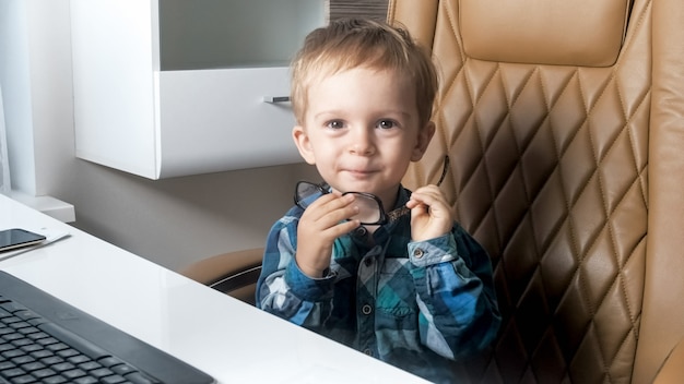 Porträt des netten lächelnden kleinen Jungen, der auf Bürostuhl hinter Schreibtisch im Büro sitzt.