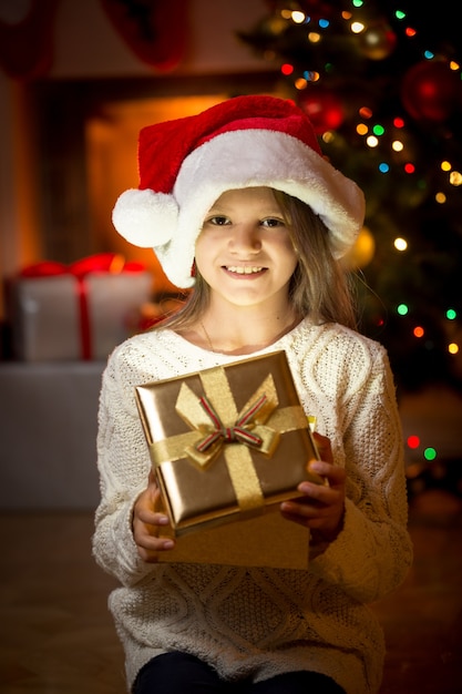 Porträt des netten lächelnden Mädchens, das gegen Kamin und Weihnachtsbaum mit Geschenkbox aufwirft