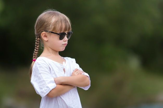 Porträt des niedlichen hübschen lustigen jungen Mädchens mit den blonden Zöpfen im weißen Kleid und in der dunklen Sonnenbrille.