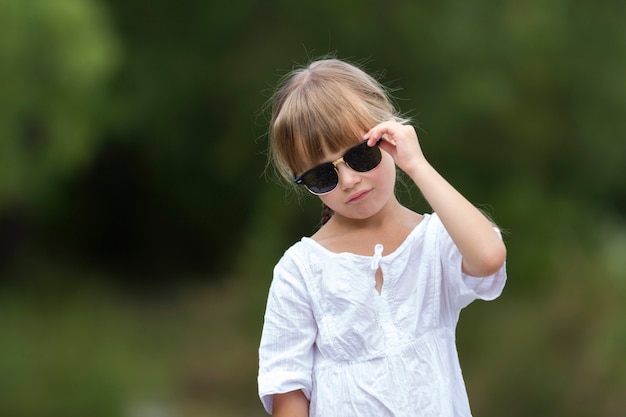 Porträt des niedlichen hübschen lustigen jungen Mädchens mit den blonden Zöpfen im weißen Kleid und in der dunklen Sonnenbrille.