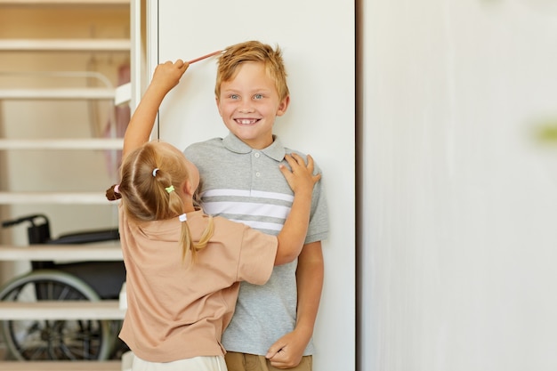 Porträt des niedlichen Mädchens, das Höhe des glücklichen älteren Bruders misst, der gegen weiße Wand zu Hause steht, kopiert Raum