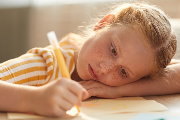 Porträt des niedlichen rothaarigen Mädchens, das Bilder zeichnet, während sie ihren Kopf im Sonnenlicht auf Tisch legt