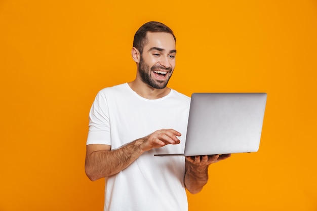 Porträt des optimistischen Mannes 30s im weißen T-Shirt, das silbernen Laptop hält, lokalisiert