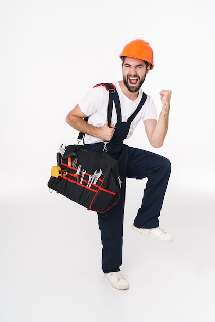 Porträt des positiv aufgeregten jungen Baumeisters im Helm lokalisiert über der weißen Wand, die Tasche mit Ausrüstungsinstrumenten hält, machen Siegergeste.