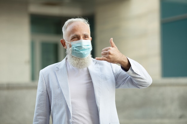 Porträt des positiven gutaussehenden älteren Geschäftsmannes mit der medizinischen Maske, die freundlich lächelt und Hand winkt