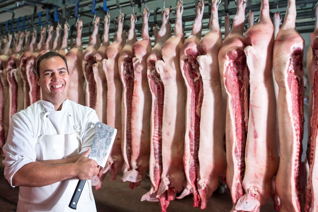 Porträt des professionellen Metzgers im Fabrikkühlraum, der Fleischhacker mit Schweineschlachtkörper hält