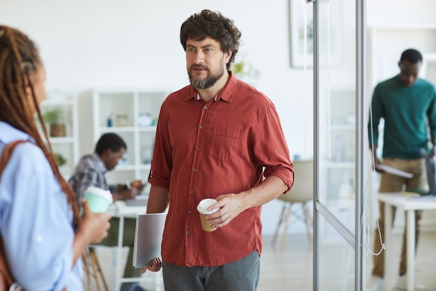 Porträt des reifen bärtigen Mannes gekleidet in der Freizeitkleidung, die mit Kollegin während der Kaffeepause im Büro spricht