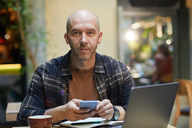 Porträt des reifen Mannes, der Handy verwendet und beim Sitzen am Tisch vor Laptop im Café schaut