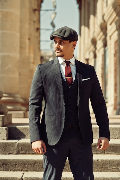Porträt des Retro-englischen Geschäftsmanns der 1920er Jahre, der dunklen Anzug, Krawatte und flache Kappe nahe alten Säulen trägt.
