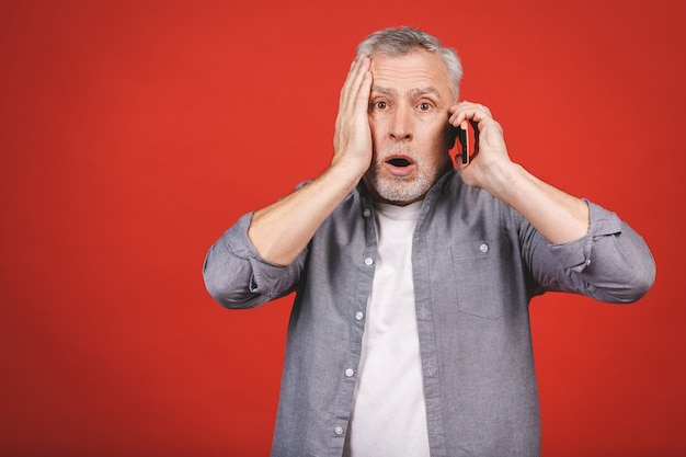 Porträt des schockierten älteren Mannes im lässigen keuchenden öffnenden Mund von der Sorge und Überraschung, die Handy hält und in der Kamera lokalisiert über rote Wand schaut.
