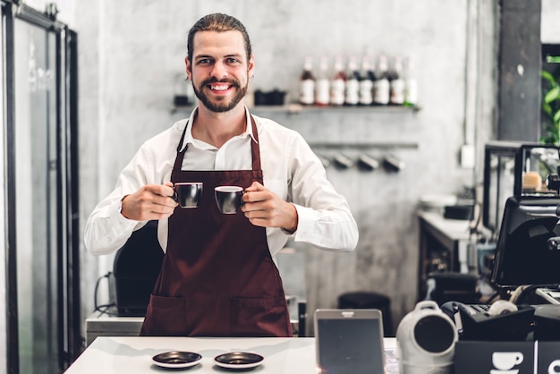 Porträt des schönen bärtigen Barista-Mannes Kleinunternehmer, der lächelt und Tasse Kaffee im Café oder im Café hält. Männlicher Barista, der am Café steht