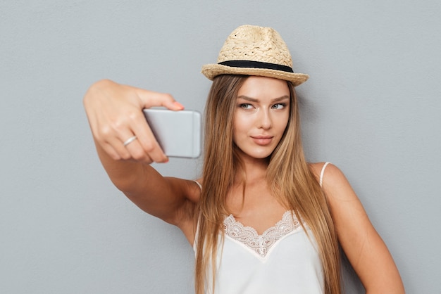 Porträt des schönen jungen Mädchens im Hut, das selfie mit Smartphone lokalisiert auf einem grauen Hintergrund macht