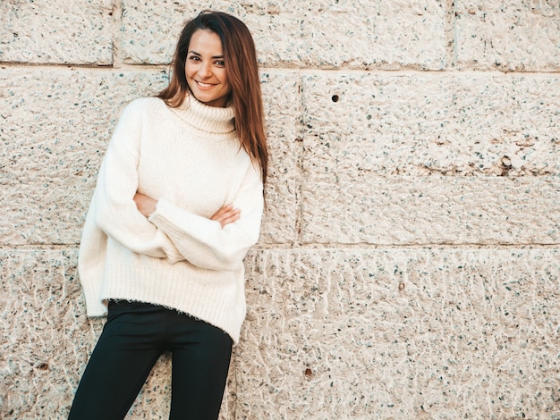 Porträt des schönen lächelnden Baumusters. Frau gekleidet in warmen Hipster-weißen Pullover. Posiert in der Nähe der Wand auf der Straße