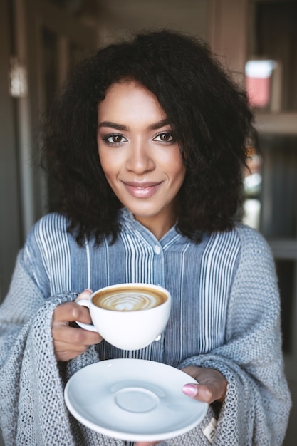 Porträt des schönen Mädchens mit dem dunklen lockigen Haar, das im Plaid mit Tasse in den Händen eingewickelt wird