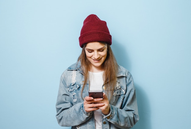 Porträt des schönen schreienden jungen Mädchens, welches den Smartphone zeigt Siegergeste, lokalisiert über blauem Hintergrund hält