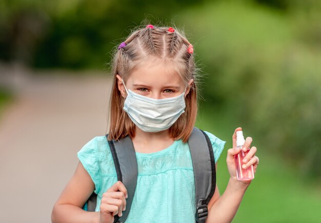 Porträt des Schulmädchens in der Maske, die Desinfektionsmittel auf Natur hält