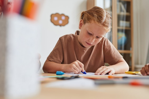 Porträt des sommersprossigen rothaarigen Mädchens, das Bilder mit Buntstiften zeichnet, während Kunstunterricht in der Schule oder im Entwicklungszentrum genießt