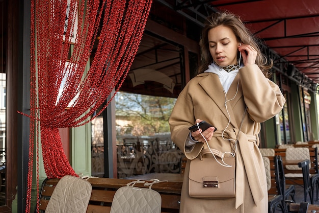 Porträt des tragenden Mantels des jungen Mädchens, der auf die Musik auf Caféveranda mit den Kopfhörern halten Handy in den Händen hört