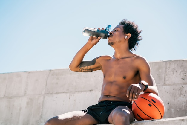 Porträt des Trinkwassers des Afroathletenmannes und des Haltens des Basketballballs im Freien. Sport und gesunder Lebensstil.