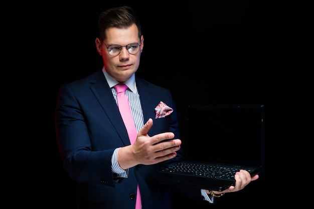 Porträt des überzeugten hübschen ernsten Geschäftsmannes, der auf dem Laptop macht das Handzeichen lokalisiert auf Schwarzem darstellt