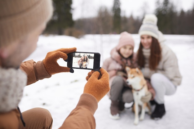 Porträt des Vaters, der ein Foto von süßer Tochter und Frau mit Hund macht, während sie gemeinsam im Winterwald spazieren gehen, sich auf den Smartphone-Bildschirm konzentrieren, Platz kopieren