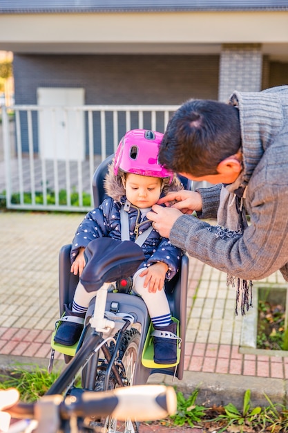 Porträt des Vaters, der ihrer kleinen Tochter, die im Fahrradsitz sitzt, einen Fahrradsicherheitshelm schließt. Sicheres und Kinderschutzkonzept.