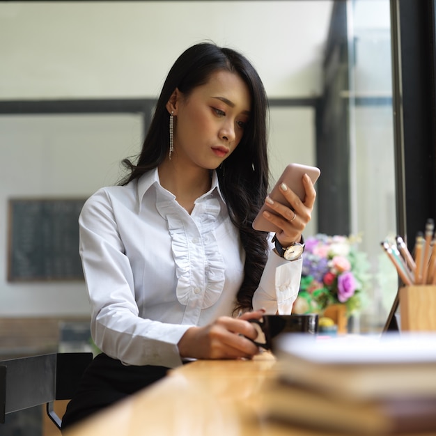 Porträt des weiblichen Büroangestellten, der am Arbeitstisch mit Kaffeetasse und Smartphone entspannt