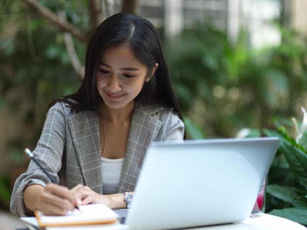 Porträt des weiblichen Schreibens auf leerem Notizbuch, während Laptop auf Couchtisch arbeiten