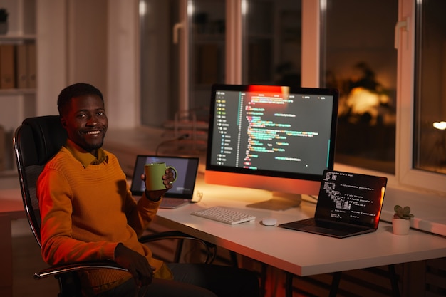 Porträt des zeitgenössischen afroamerikanischen Mannes, der Code schreibt und Kamera beim Entspannen am Arbeitsplatz mit Kaffeetasse, Kopierraum betrachtet