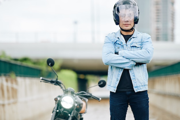 Porträt des zuversichtlichen ernsten asiatischen Mannes im Motorradhelm, der draußen mit verschränkten Armen steht