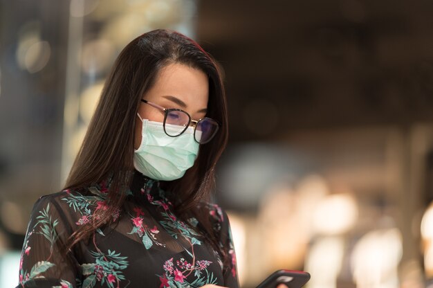 Porträt Eine schöne Frau, die mit einem Mobiltelefon steht, um E-Mail zu überprüfen und Gesichtsmaske mit Bokeh-Unschärfehintergrund zu tragen