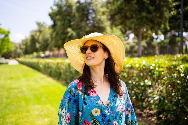 Porträt einer abgelenkten Frau mit Sonnenbrille und Strohhut, die auf einem Garten steht