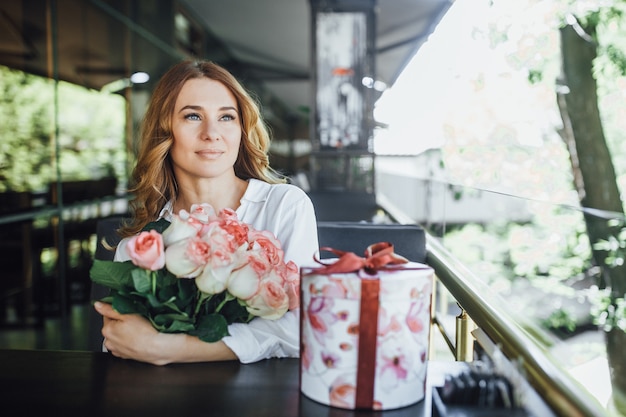 Porträt einer älteren Frau, die einen Strauß Rosen und ein Geschenk auf der Sommerterrasse eines modernen Cafés hält.