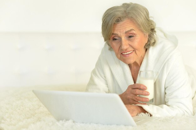 Porträt einer älteren Frau, die mit Laptop auf dem Bett liegt und Milch trinkt