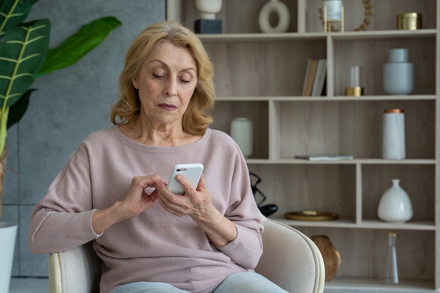 Porträt einer älteren Frau, die zu Hause auf einem Stuhl sitzt und ein Smartphone benutzt